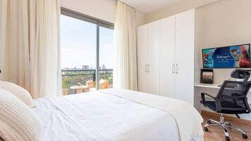 A hotel room with a large bed, a desk with a chair, a television, and a view of the city through a large window.