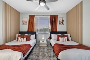A hotel room with two beds, a ceiling fan, a window with blinds, and a nightstand with a lamp.