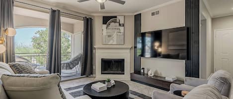 A modern living room with a fireplace, a ceiling fan, and a large flat-screen TV.