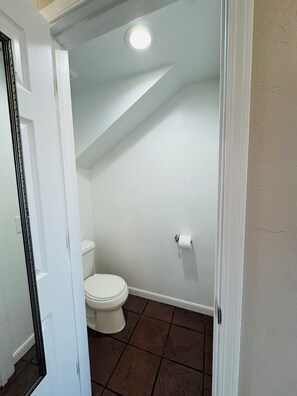 Ein kleines, schmales Badezimmer mit einer weißen Toilette, einer Toilettenpapierrolle an der Wand und braunen Fliesen auf dem Boden.
