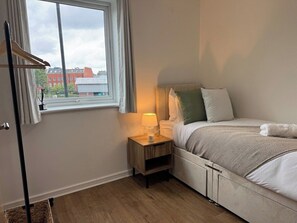 A bedroom with a bed, bedside table, lamp, and a window with a view of buildings. - Stylish Central Warrington Stay | Free Parking (Warrington)