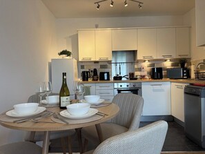 A modern kitchen with white cabinetry, a dining table set with white plates, bowls, and cutlery, and a bottle of wine. - Stylish Central Warrington Stay | Free Parking (Warrington)
