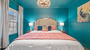 A bedroom with a large bed, two bedside tables with lamps, a chandelier, and a painting on the wall.