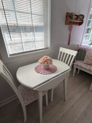 Une table à manger blanche, ornée d’un chemin de table en crochet et d’un vase de fleurs. Des chaises blanches à motifs floraux sont disposées autour de la table. Une fenêtre avec des stores est visible, et une étagère décorative est fixée au mur.