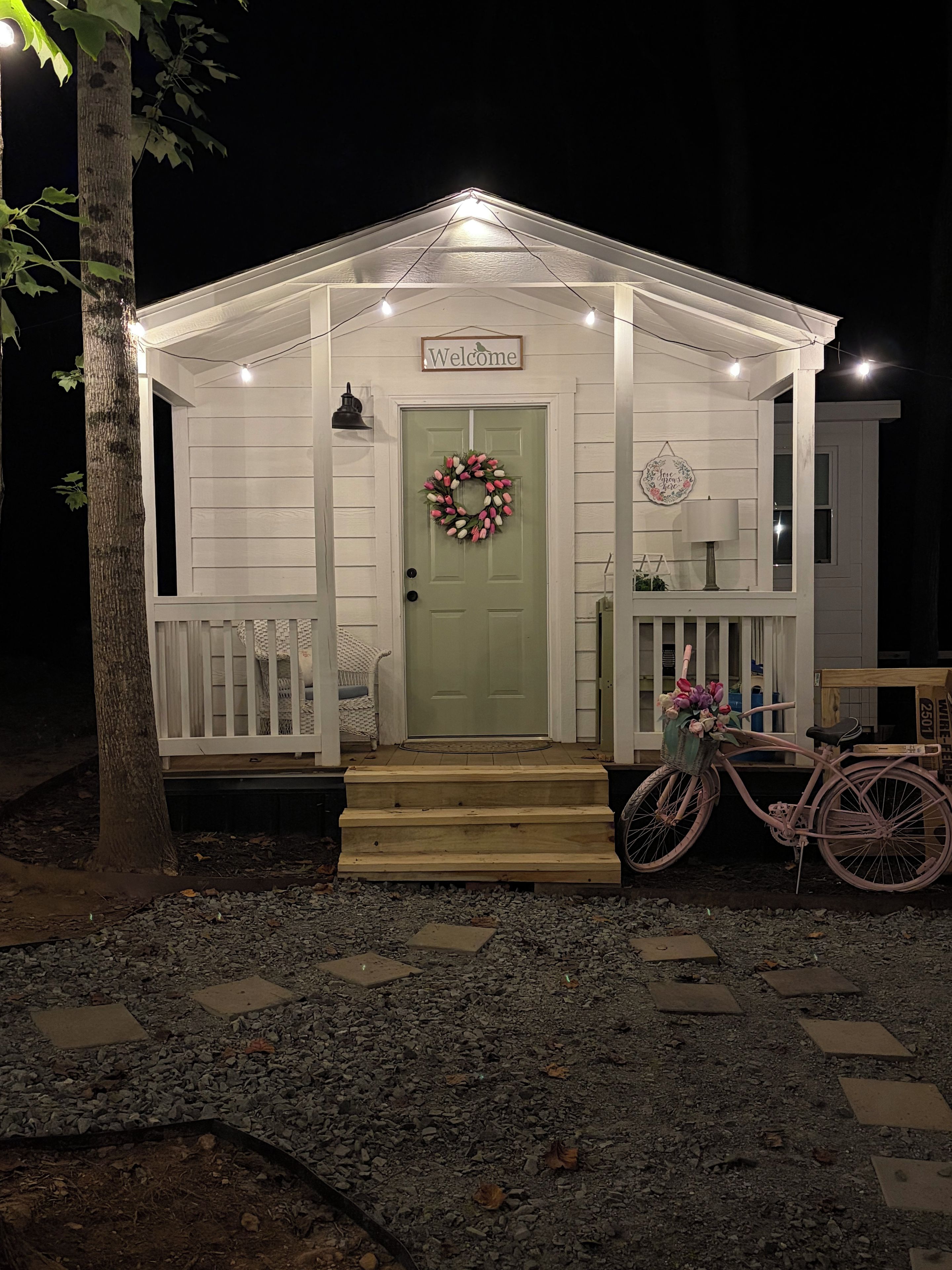 A small, white, single-story building with a green door, a welcome sign, a wreath, and a bicycle with flowers.