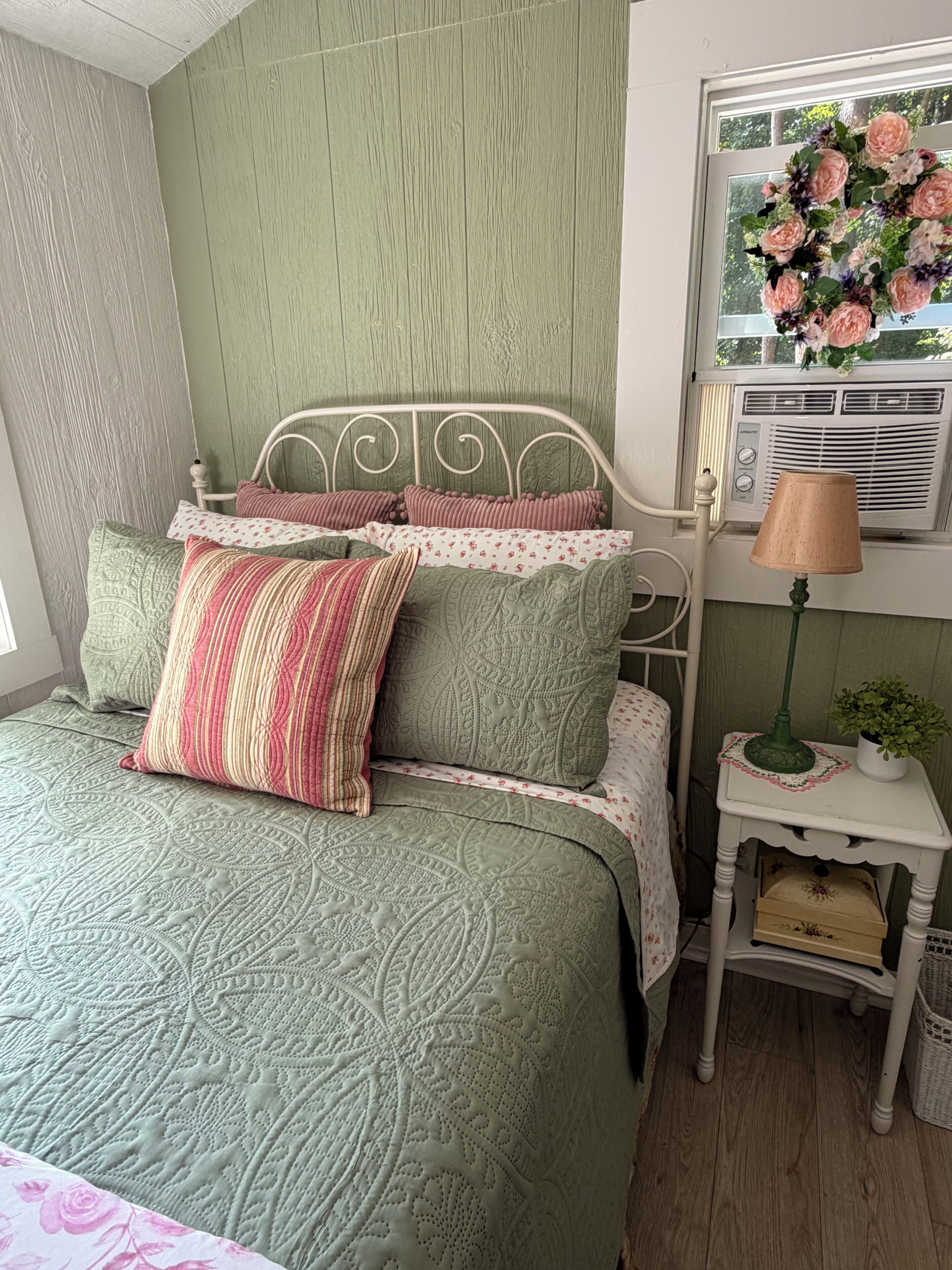 A bed with a green quilt, multiple pillows, a bedside table with a lamp, and a window with a floral decoration.
