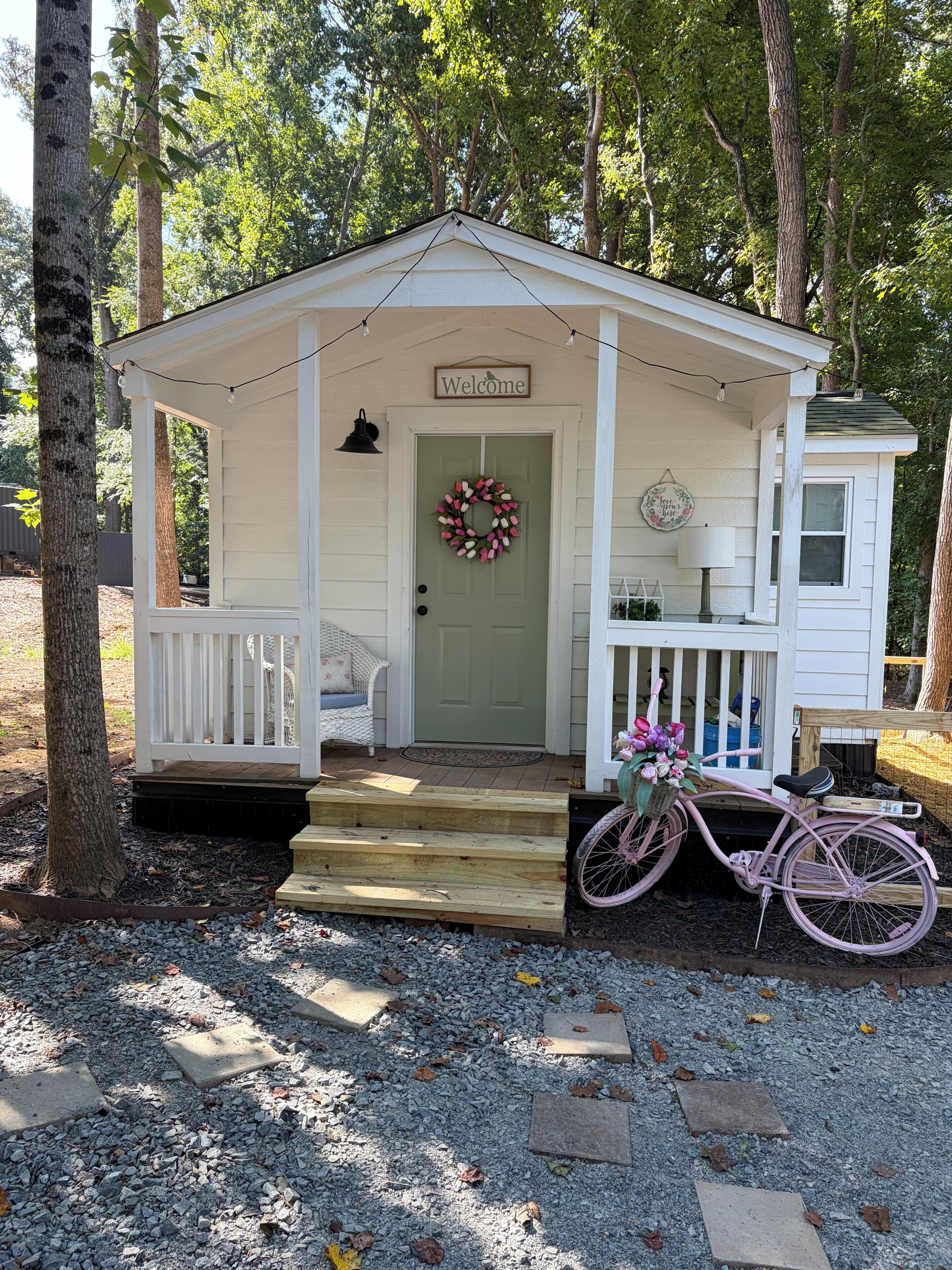 Une petite cabane blanche avec un porche, une pancarte d’accueil et un vélo décoré de fleurs.