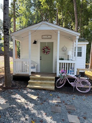 Une petite cabane blanche avec un porche, une pancarte d’accueil et un vélo décoré de fleurs.