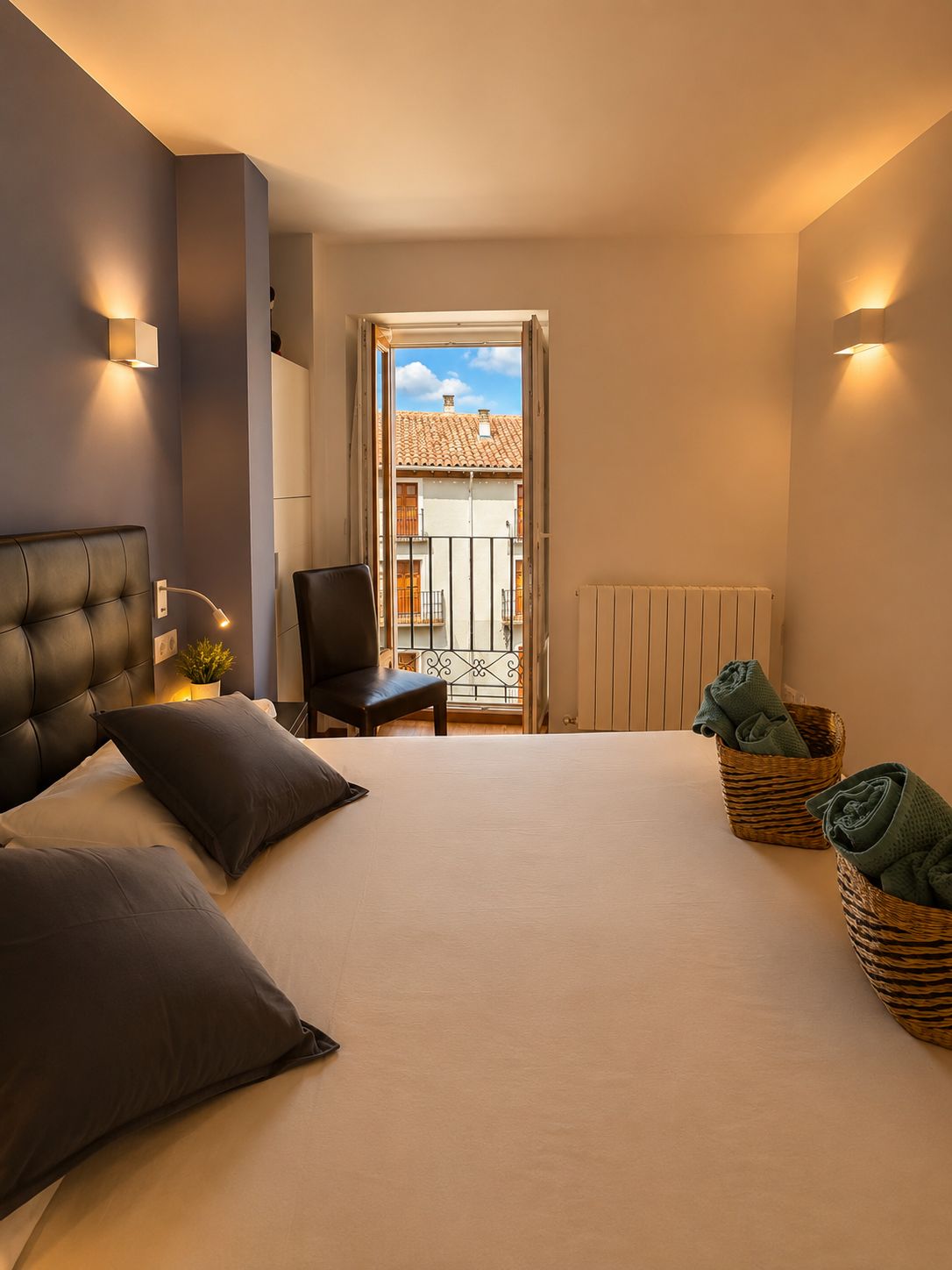 Une chambre à coucher comprenant un lit, une chaise, une petite table, un radiateur et une vue sur un balcon doté d’une balustrade.