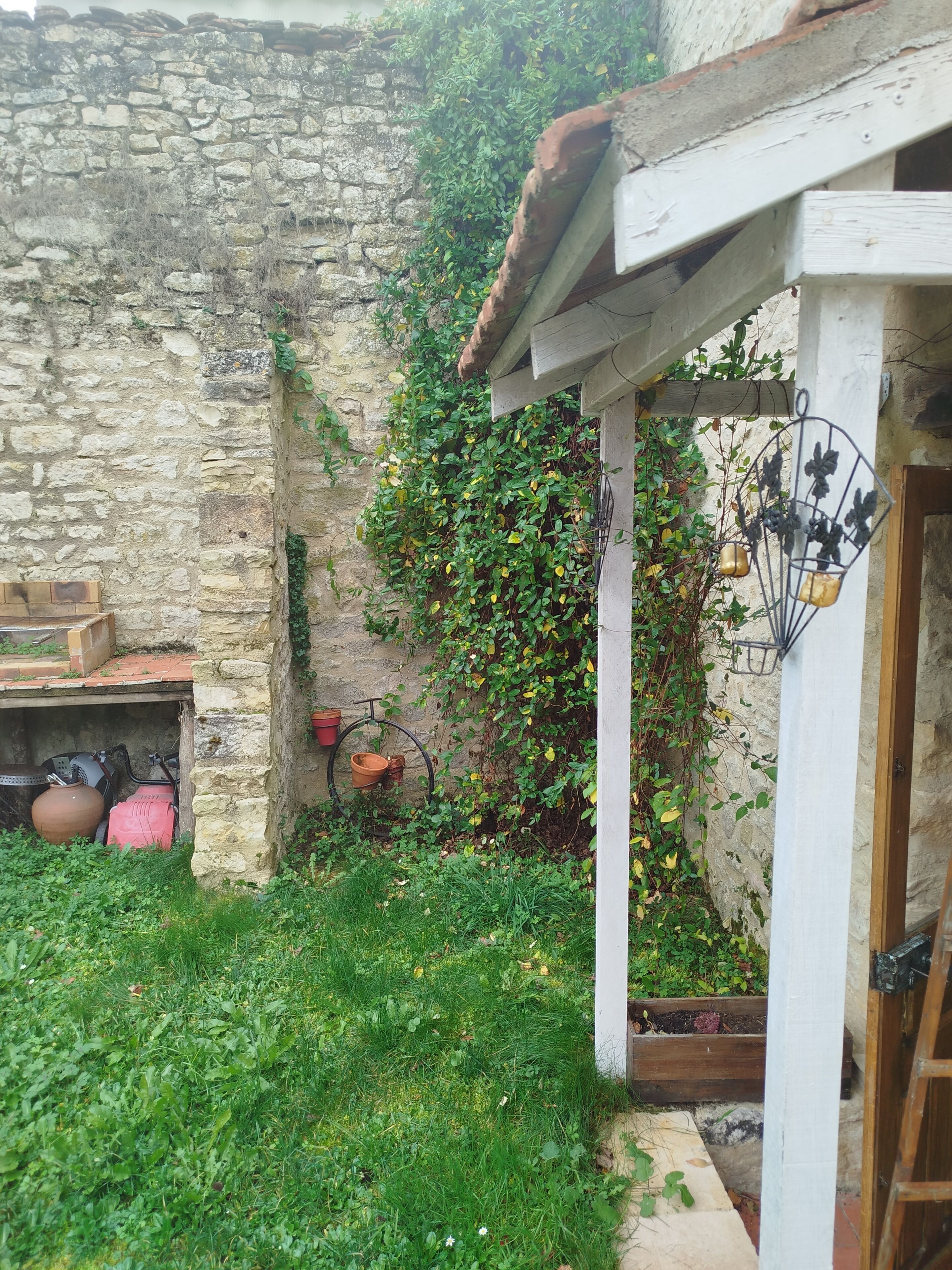 Un mur de pedra rústic amb una porta de fusta, una petita zona de jardí amb gespa i plantes, i una bicicleta adossada a la paret.