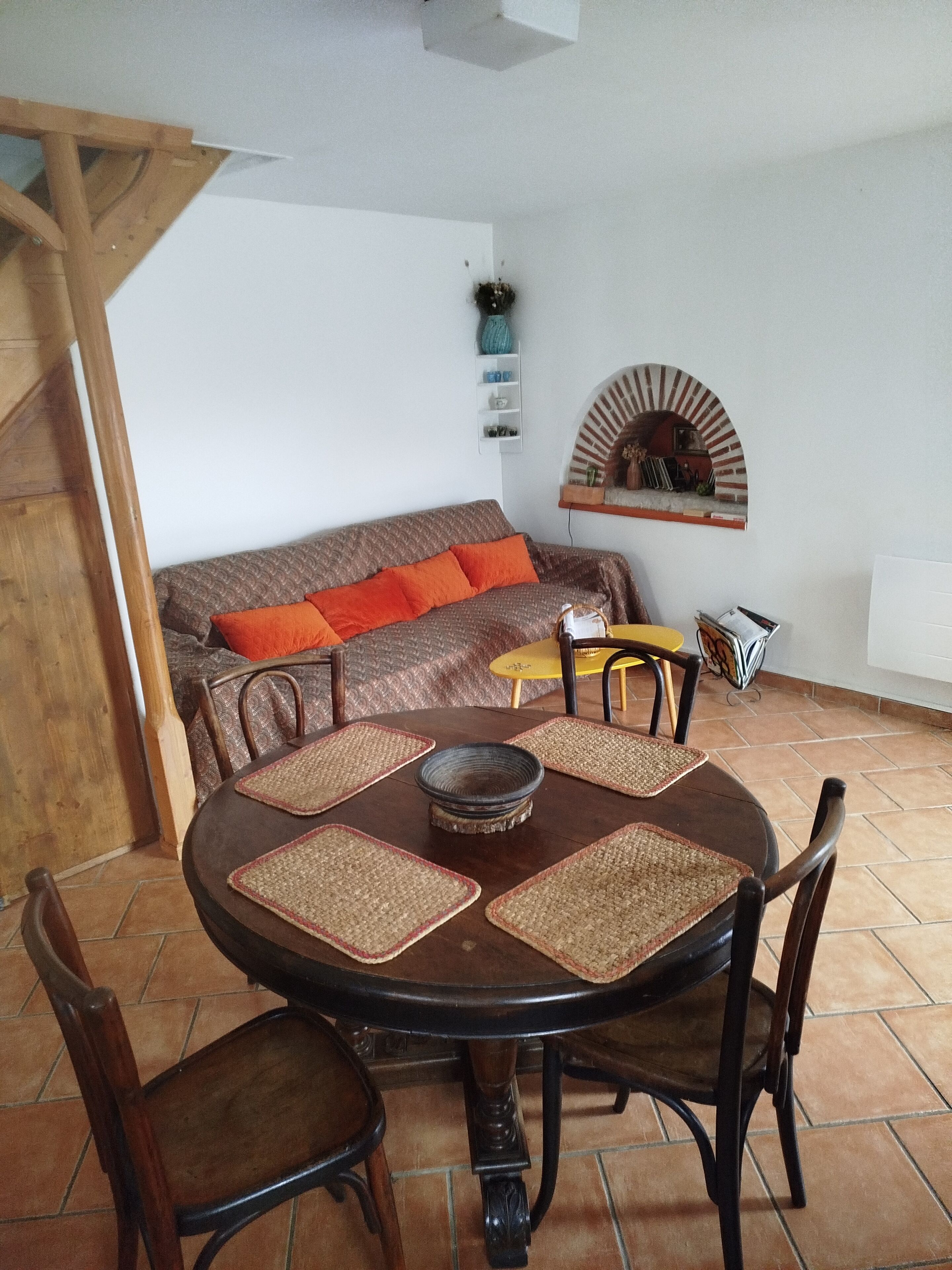 Een eethoek met een ronde tafel, stoelen en een bank met oranje kussens.