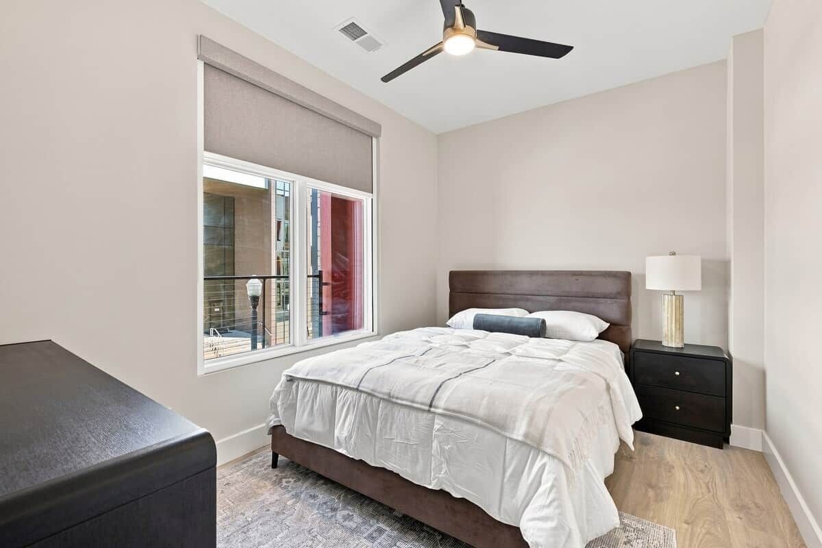 Une chambre à coucher comprenant un lit, une table de chevet, une lampe, un ventilateur de plafond et une fenêtre donnant sur des immeubles.