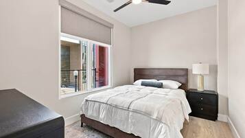 Une chambre à coucher comprenant un lit, une table de chevet, une lampe, un ventilateur de plafond et une fenêtre donnant sur des immeubles.