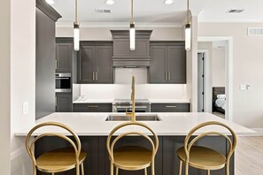 A modern kitchen with dark cabinets, a central island with a sink, and three wooden chairs. - The Regent · 2 Bed · 2 Bath · 1,300+ SF (Owensboro)