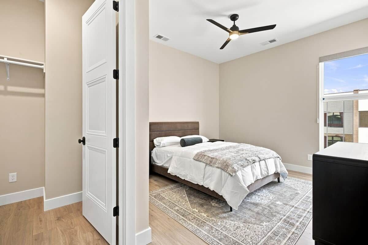 Une chambre avec un lit, un ventilateur de plafond, une porte blanche et une vue sur l’extérieur.