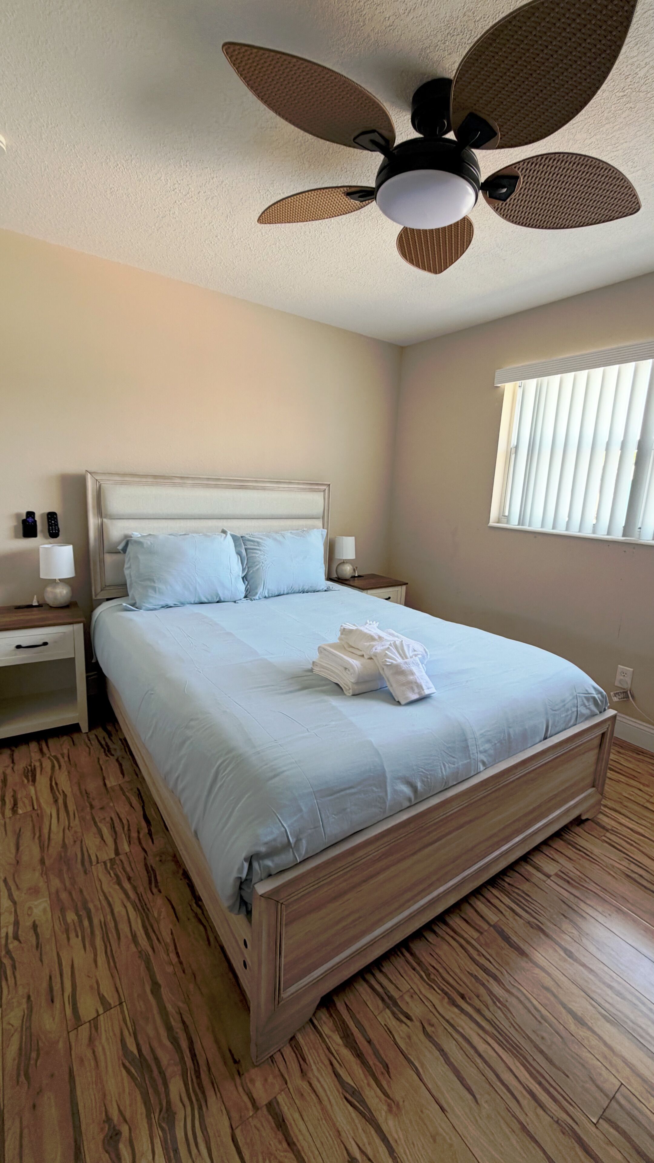 Une chambre à coucher comprenant un lit, des tables de chevet, un ventilateur de plafond et une fenêtre avec des stores.