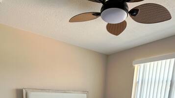 Une chambre à coucher comprenant un lit, des tables de chevet, un ventilateur de plafond et une fenêtre avec des stores.