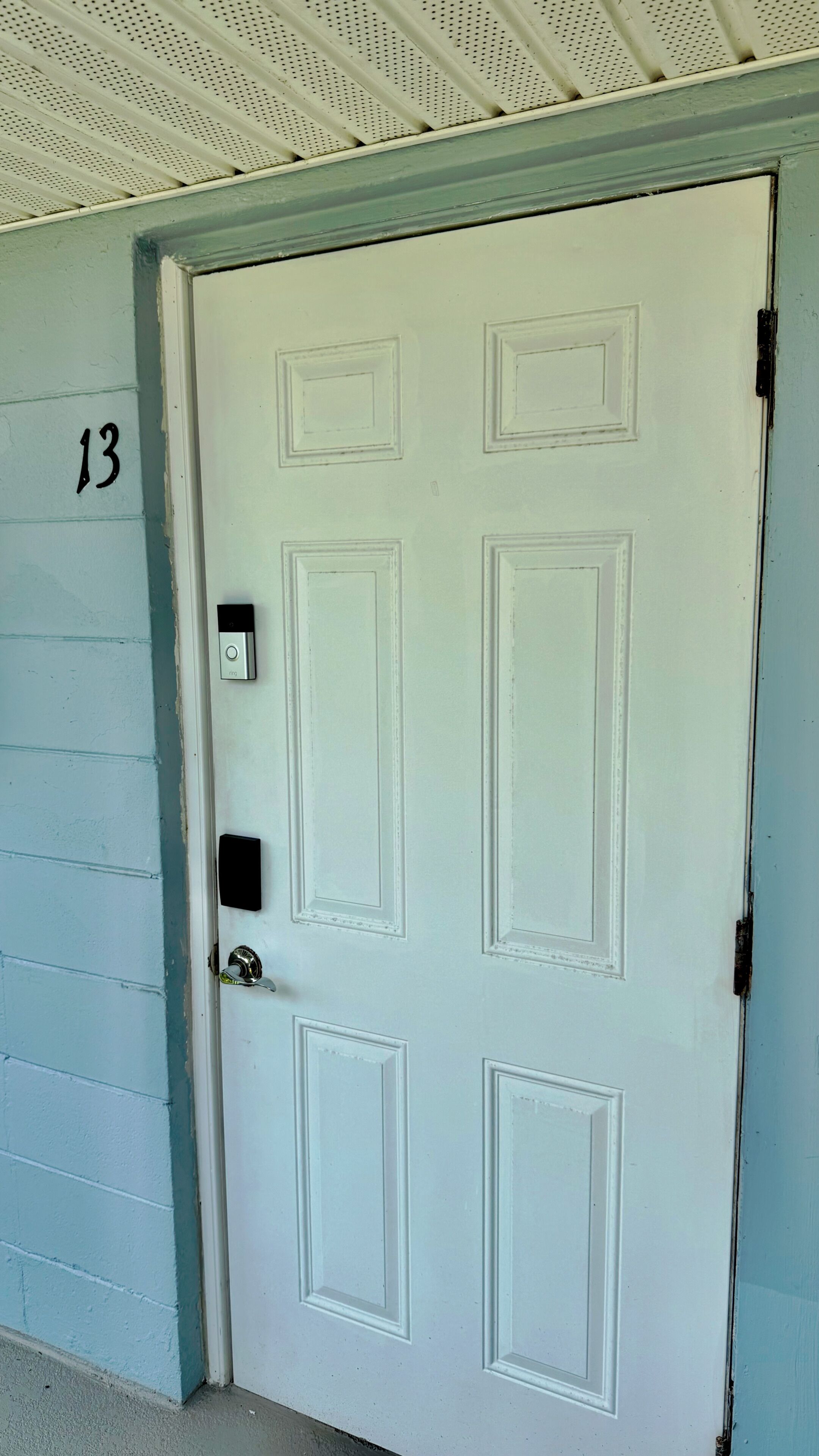Une porte blanche avec le numéro « 13 » peint sur le mur, à gauche, un interphone et une poignée de porte.