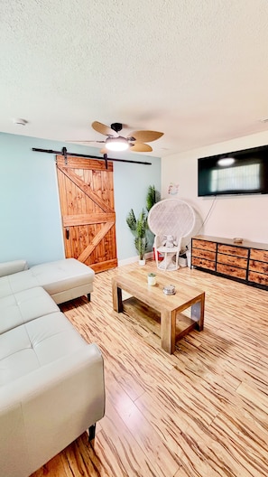 A living room with a sofa, coffee table, wooden floor, ceiling fan, and a TV mounted on the wall. - 3 Min To Beach-Sleeps 7-Close To Port & NASA-Play or Work Cape Canaveral Condo (Cape Canaveral)