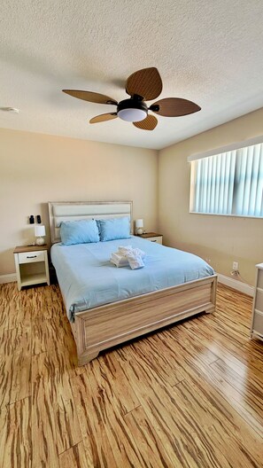 A bedroom with a bed, nightstands, a ceiling fan, a window with blinds, and a wooden floor. - 3 Min To Beach-Sleeps 7-Close To Port & NASA-Play or Work Cape Canaveral Condo (Cape Canaveral)