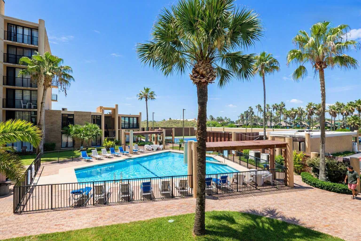 A hotel with a swimming pool, palm trees, and a clear blue sky.