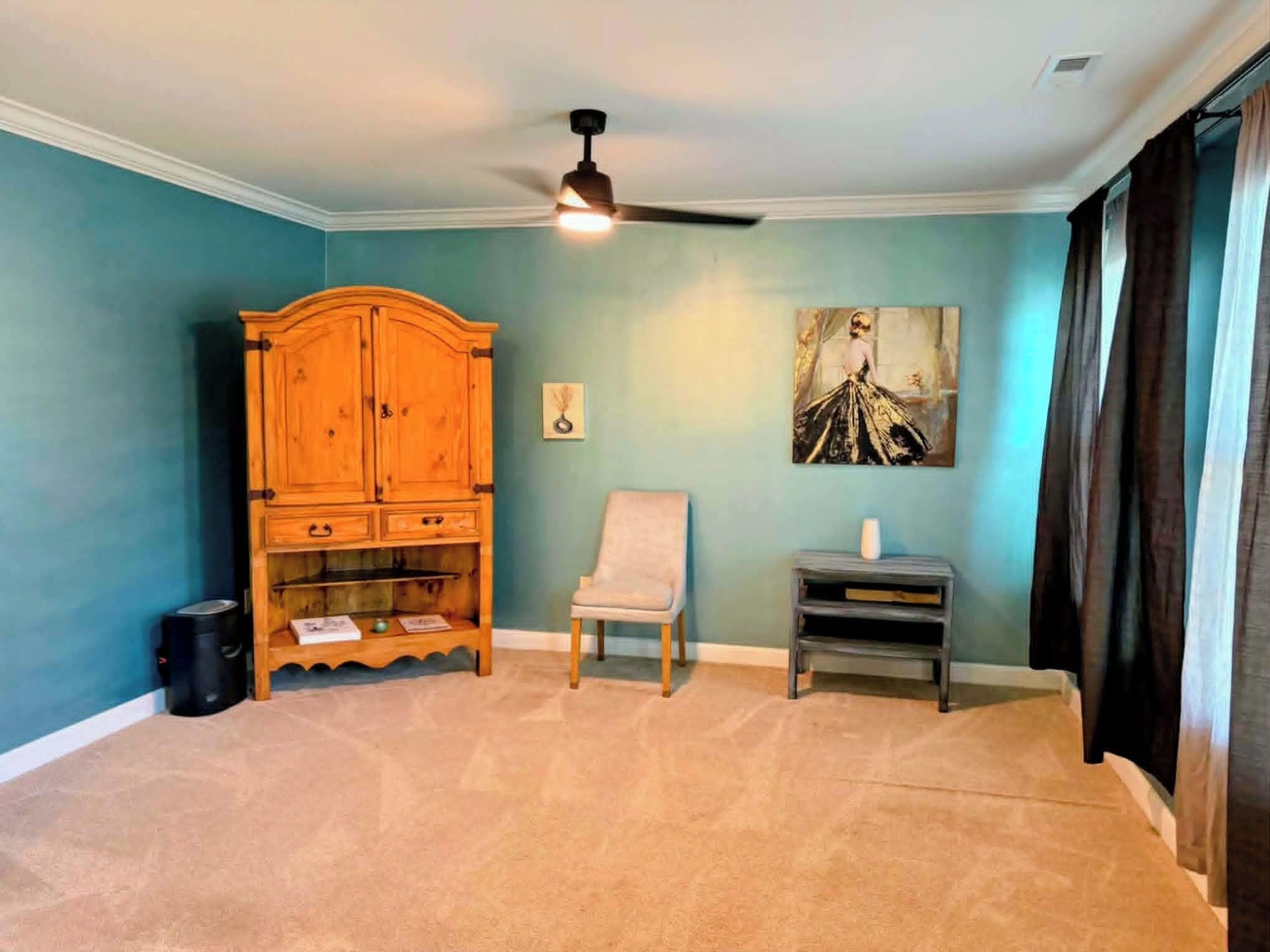 A room with a ceiling fan, a wooden cabinet, a chair, and a painting on the wall.
