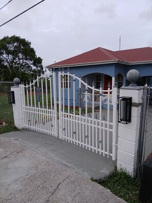 A gated entrance with a white fence and a red-roofed building in the background. - Serene 2-bedroom house with AC in Cassada Gardens (Cassada Gardens)