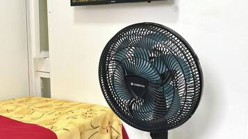 Una habitación con una cama, un ventilador de pie y un televisor con una imagen de paisaje natural.
