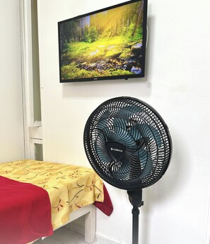 A room with a bed, a standing fan, and a mounted television displaying a nature scene. - Apartamentos RC Hospedagem (Recife)