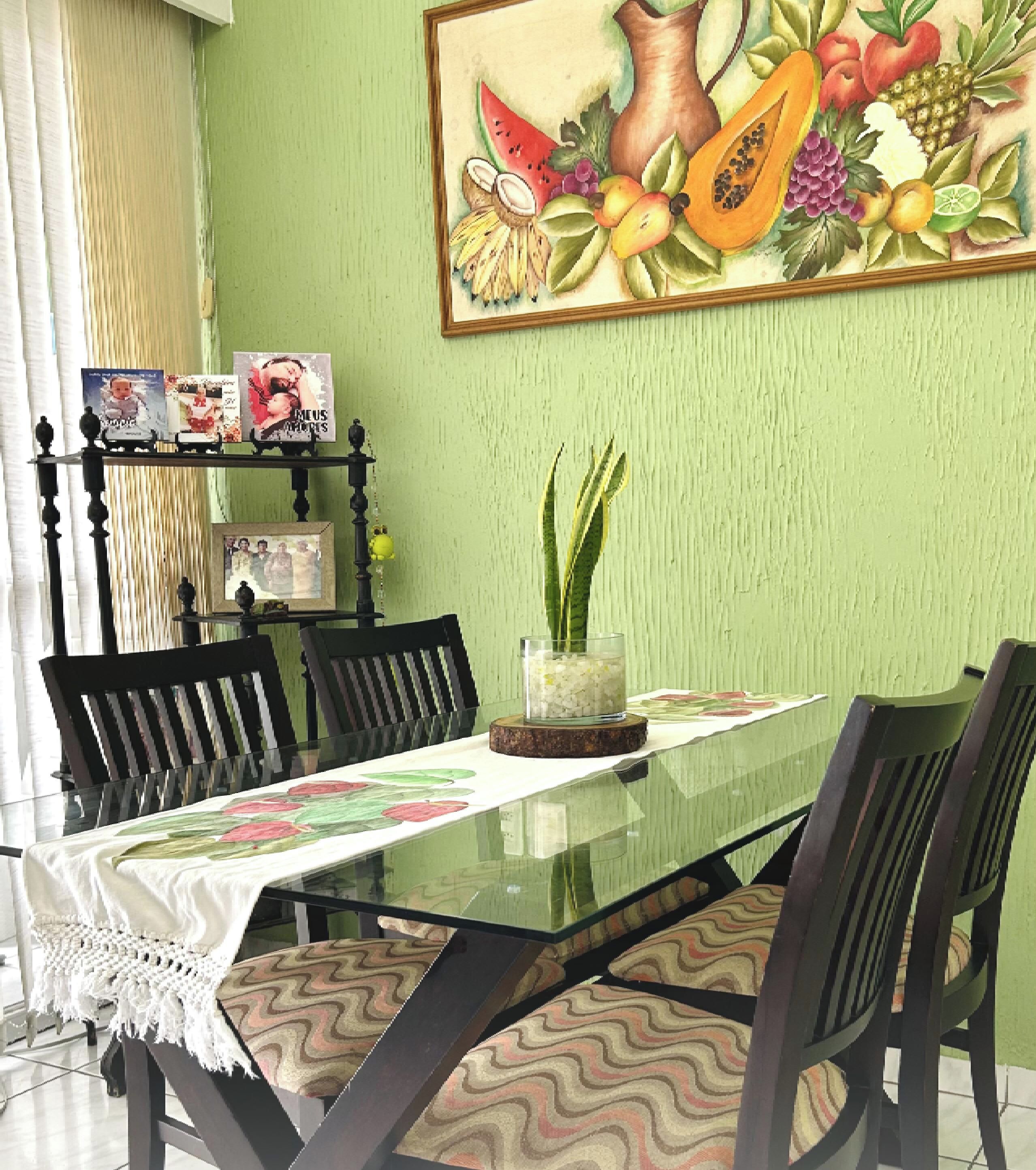 Un comedor con una mesa de vidrio, sillas y un cuadro de bodegón con frutas en la pared.