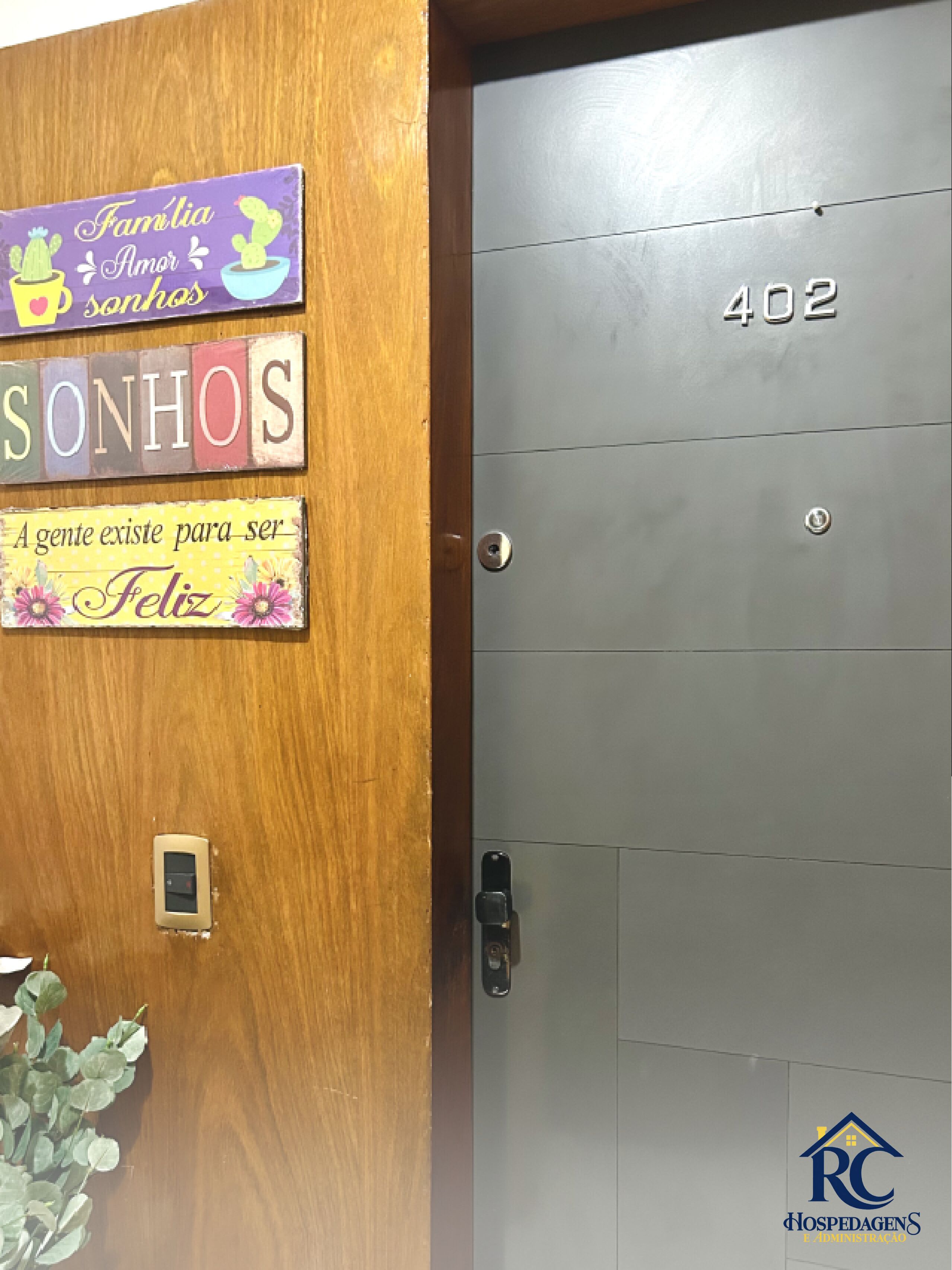 A wooden door with a sign that reads 'Família Amor Sonhos' and a metal door with the number '402' and a logo for 'HOSPEDAGENS E ADMINISTRAÇÃO'.