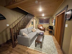 A cozy living room with a wooden staircase, a grey sofa, a leather armchair, a coffee table, and a patterned rug.
