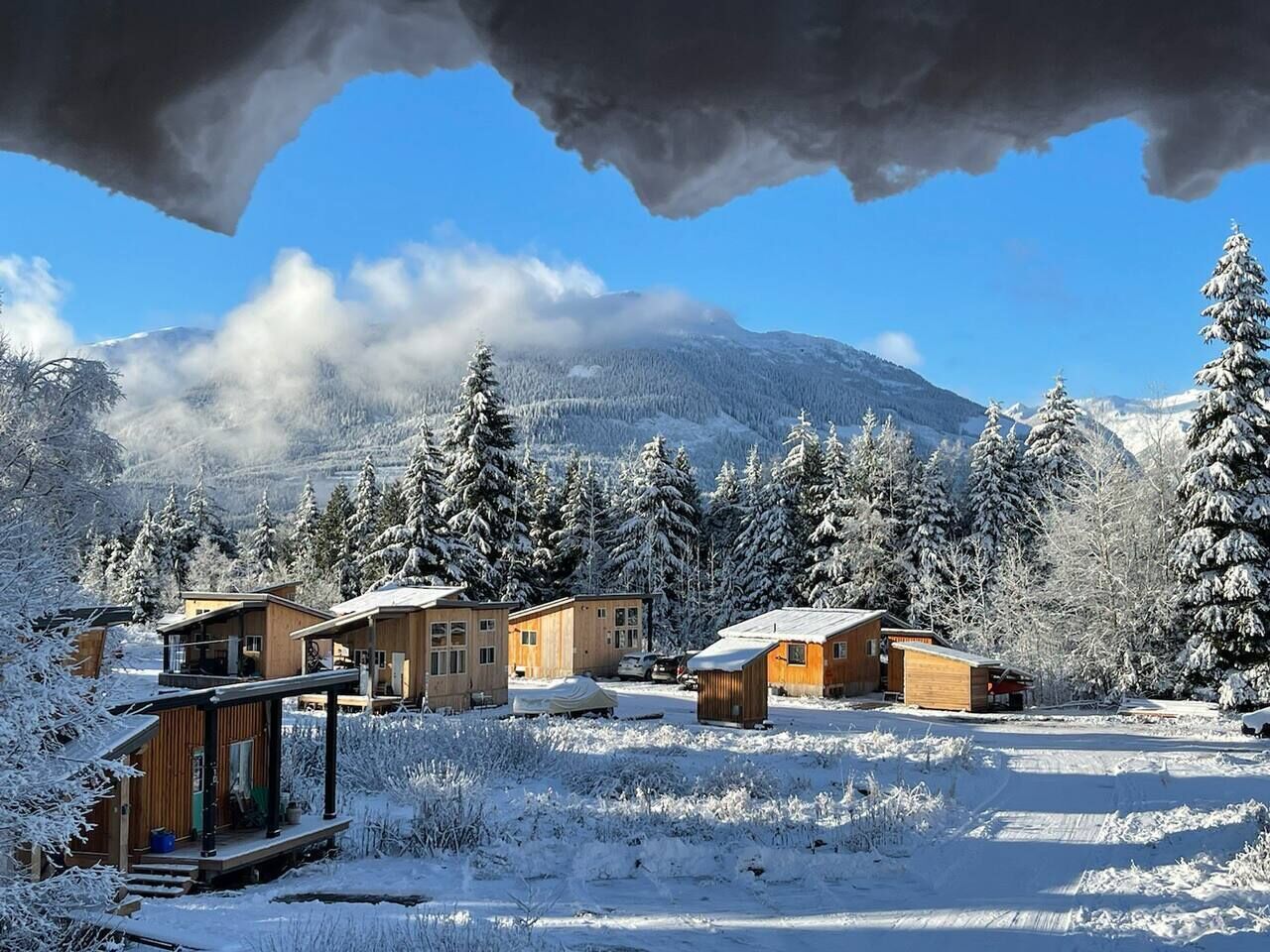 Paisaje nevado con varias cabañas de madera y una pequeña estructura de madera, probablemente una posada, con montañas al fondo.