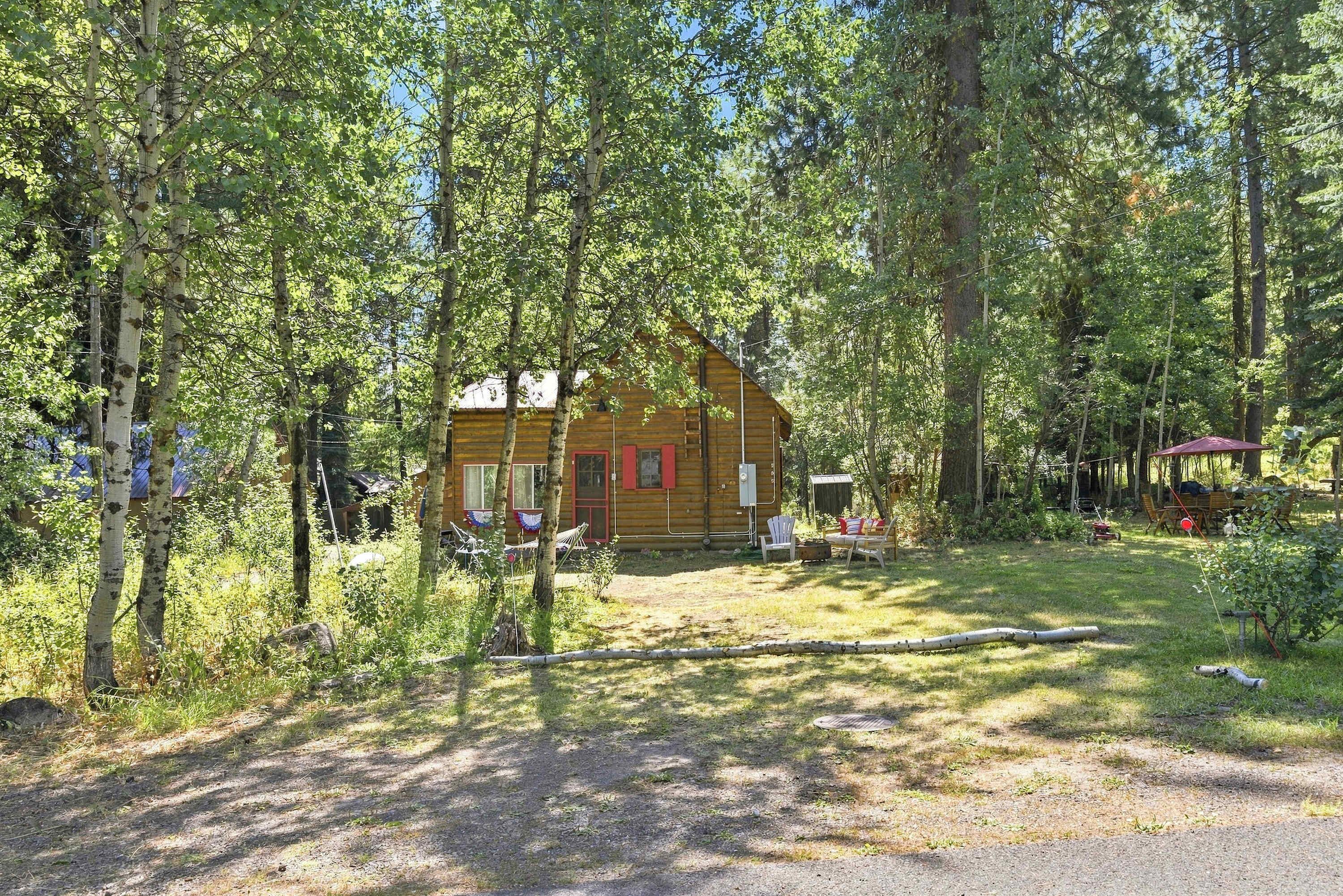 Une cabane en bois entourée d’arbres, avec une cour gazonnée et un espace extérieur aménagé pour s’asseoir.