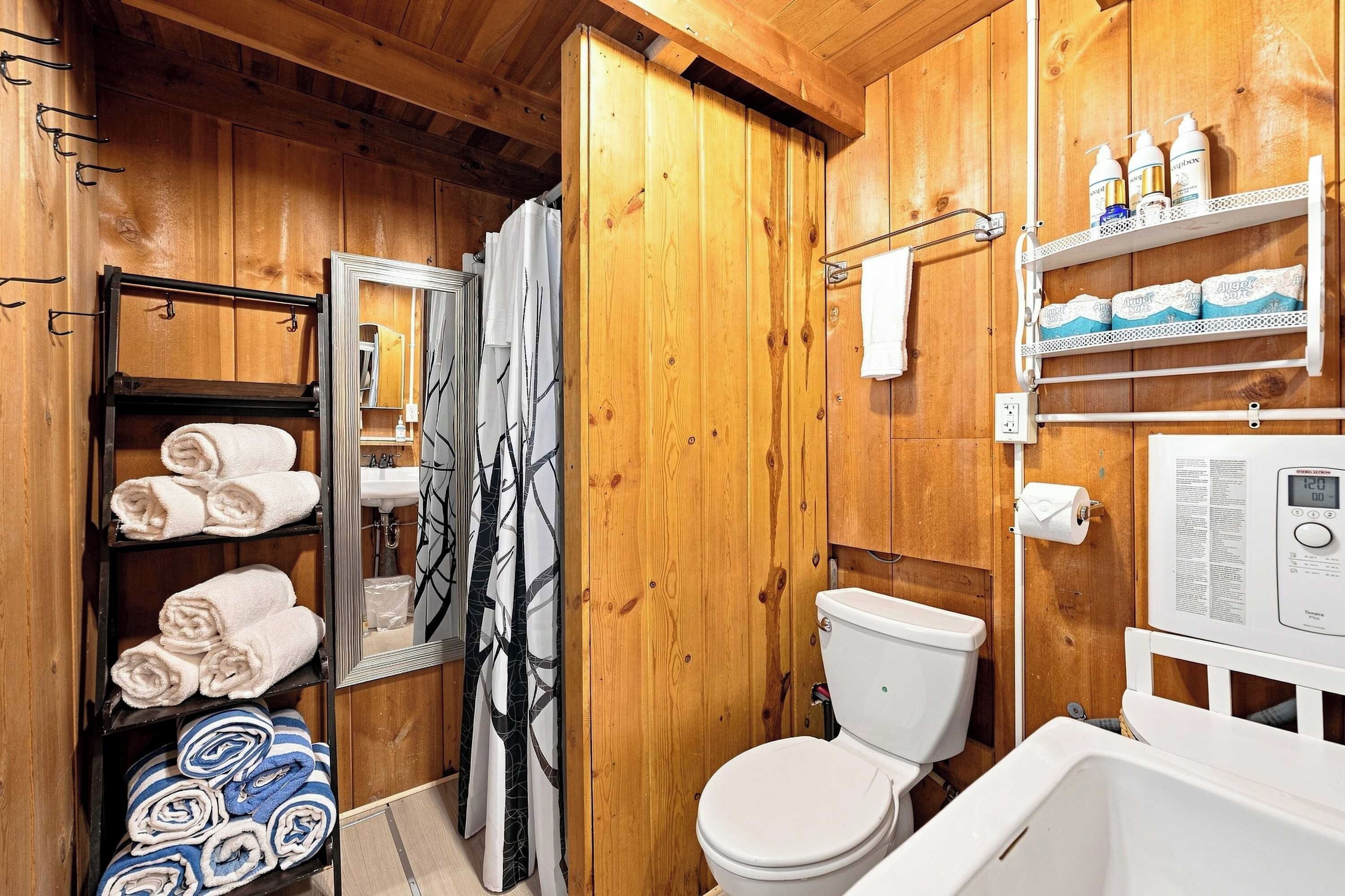 Un baño de madera con inodoro, cortina de ducha, lavamanos y estantes con artículos de aseo.