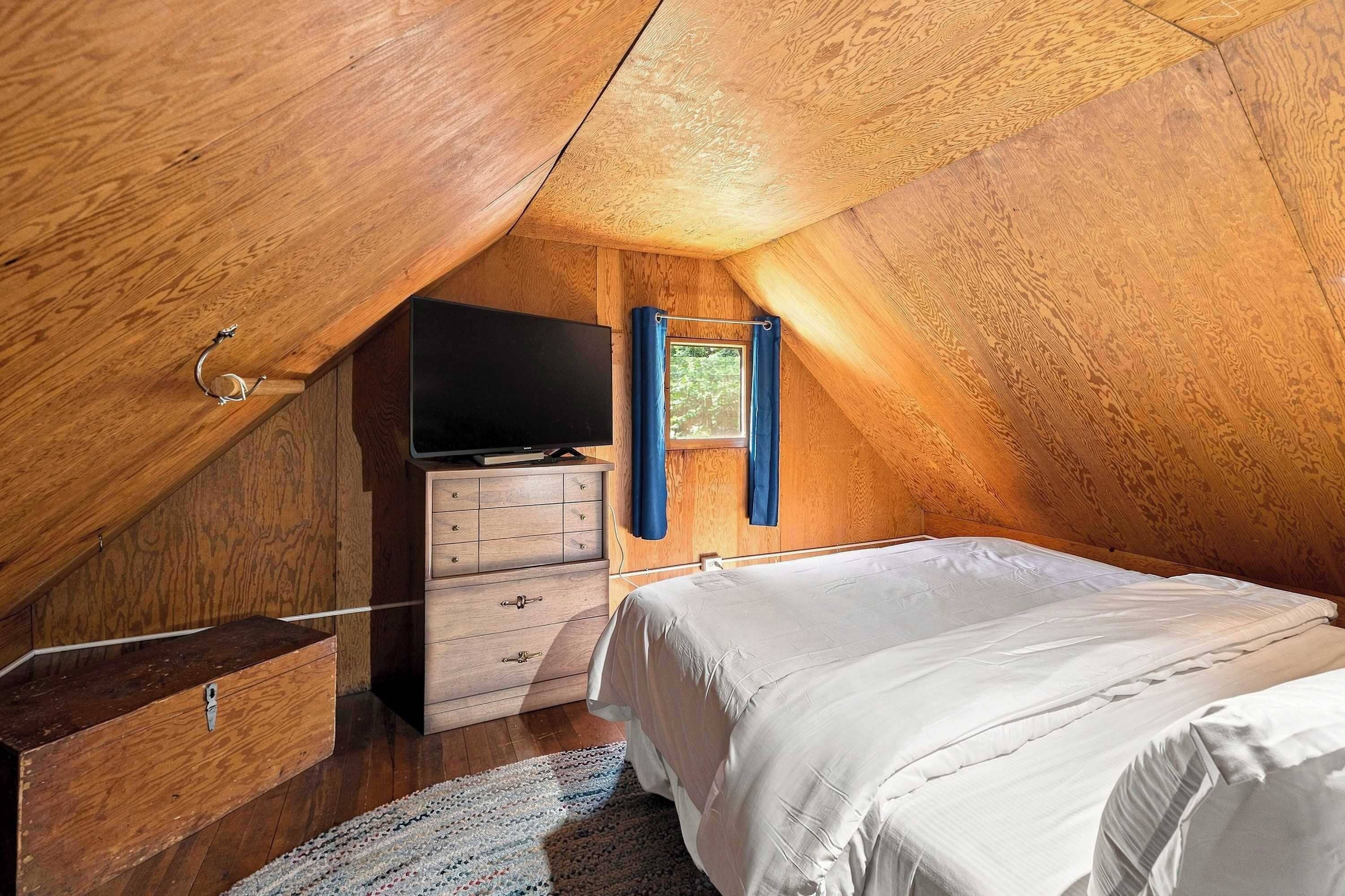 A cozy attic bedroom with a wooden sloped ceiling, a flat-screen TV, a dresser, and a bed.
