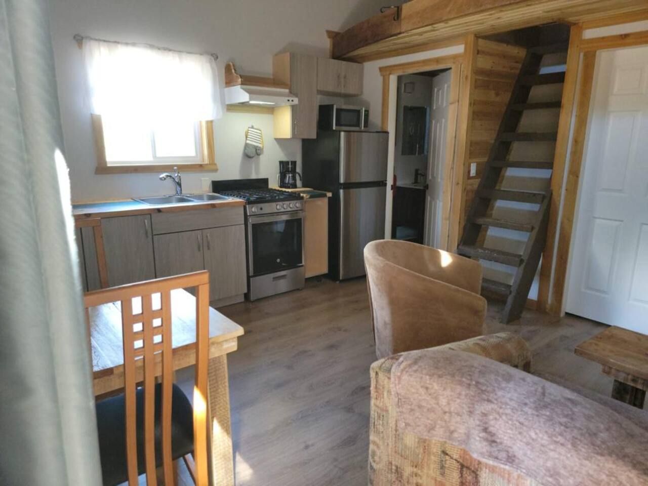 A compact kitchen with a stove, microwave, and refrigerator. There is a wooden chair and a small table. A wooden staircase is visible in the background.