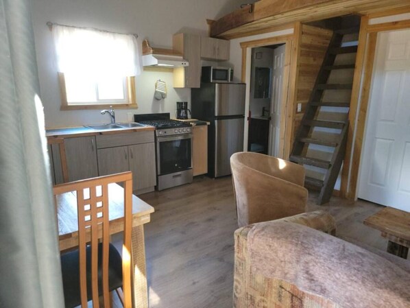 A compact kitchen with a stove, microwave, and refrigerator. There is a wooden chair and a small table. A wooden staircase is visible in the background.