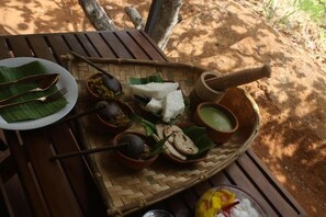 A wooden table with a woven basket containing food items, utensils, and a mortar. - Wilpattu Wilderness Camping - Deluxe Double Room -2 Adults + 1 Child- (Wilpattu)