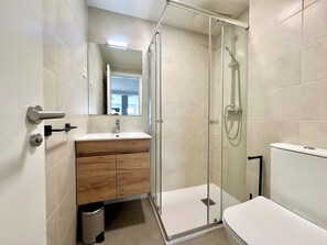 Un bany modern amb dutxa d'estuc de vidre, un tocador de fusta i un vàter blanc.