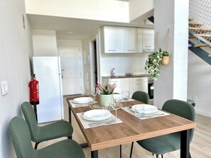 Zona de menjador amb taula de fusta preparada per a quatre, cadires tapissades de verd i una cuina al fons.