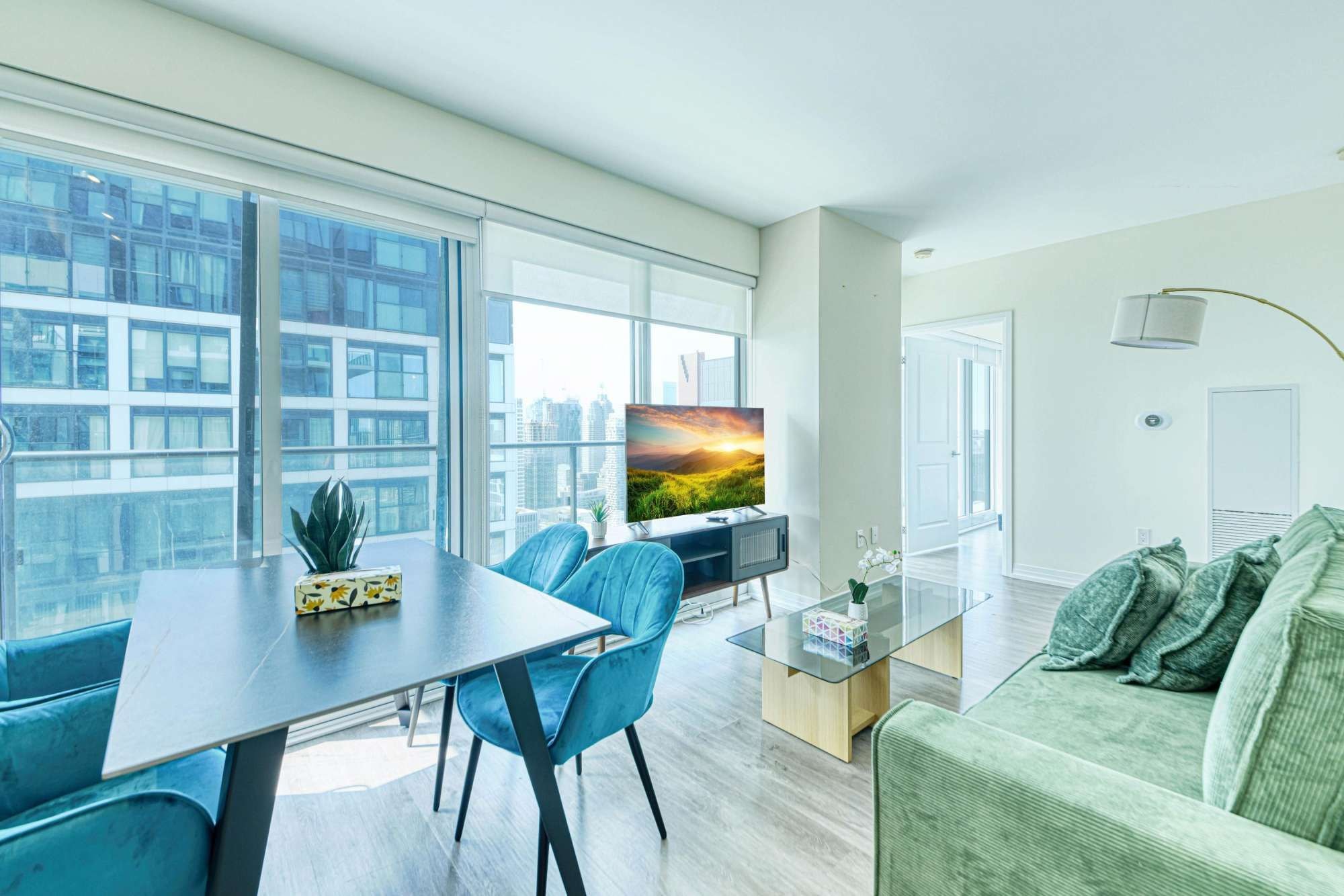 A modern living room with a dining area, large windows, a flat-screen TV, and a green sofa.