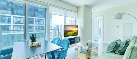 A modern living room with a dining area, large windows, a flat-screen TV, and a green sofa.