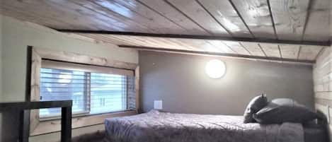 A small attic bedroom with a single bed, a window with blinds, and a wooden ceiling.