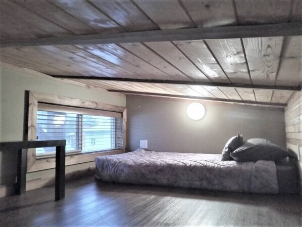 A small attic bedroom with a single bed, a window with blinds, and a wooden ceiling.