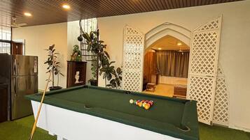 Une table de billard avec un queue de billard, entourée de gazon synthétique vert, dans une pièce dotée de panneaux de plafond en bois et d’une cloison blanche ornée d’un motif en treillis.