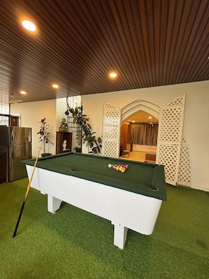 Een pooltafel met een keu, omringd door groene kunstmatige grasmat, in een ruimte met houten plafondpanelen en een witte scheidingswand met een rasterpatroon.
