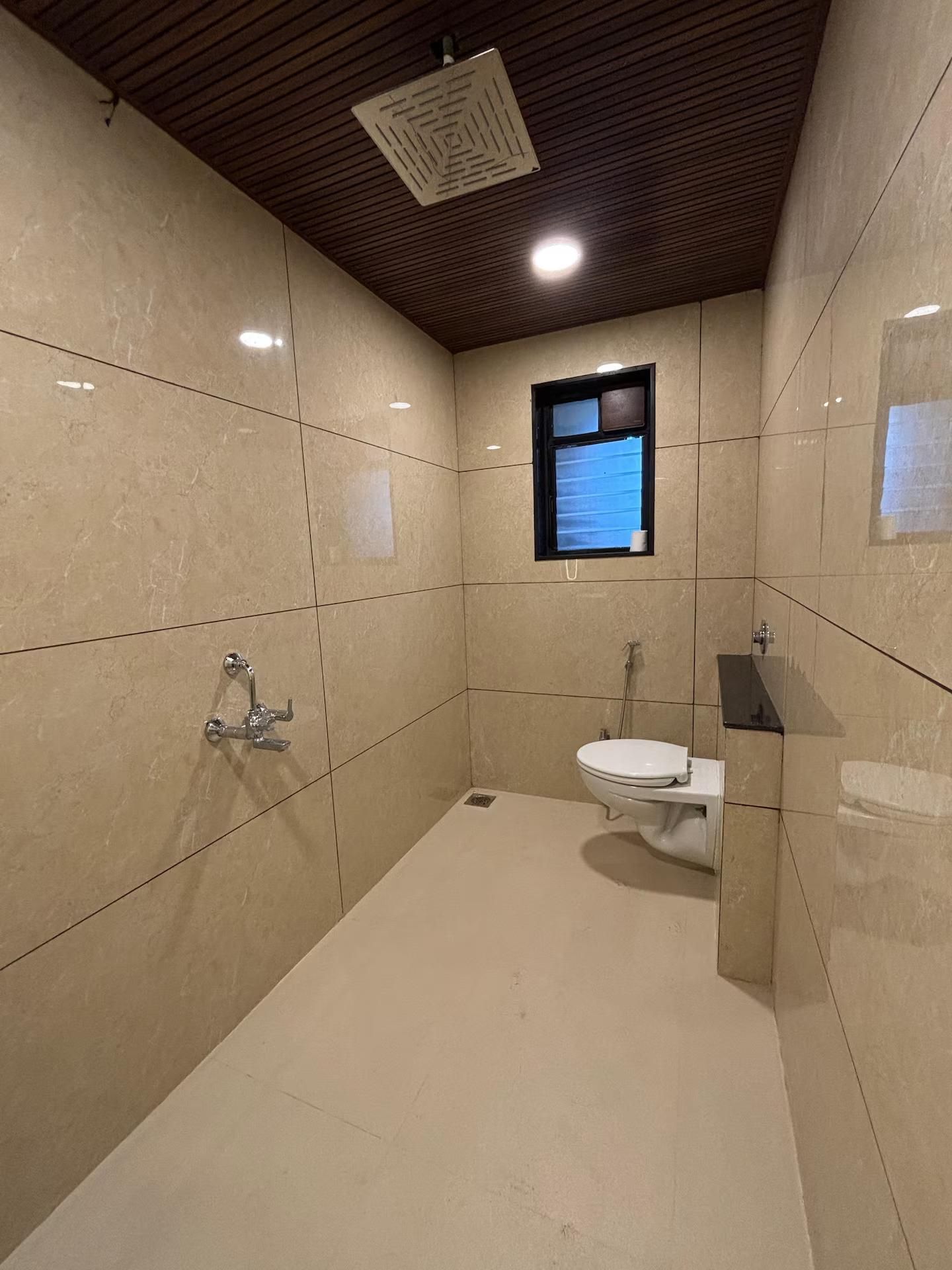 A bathroom with a shower, toilet, and sink, tiled in a light color, with a wooden ceiling and a small window.
