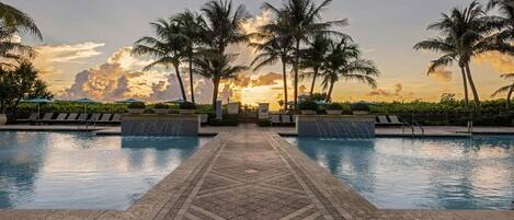 A pool area with a tiled walkway, lounge chairs, palm trees, and a sunset view.