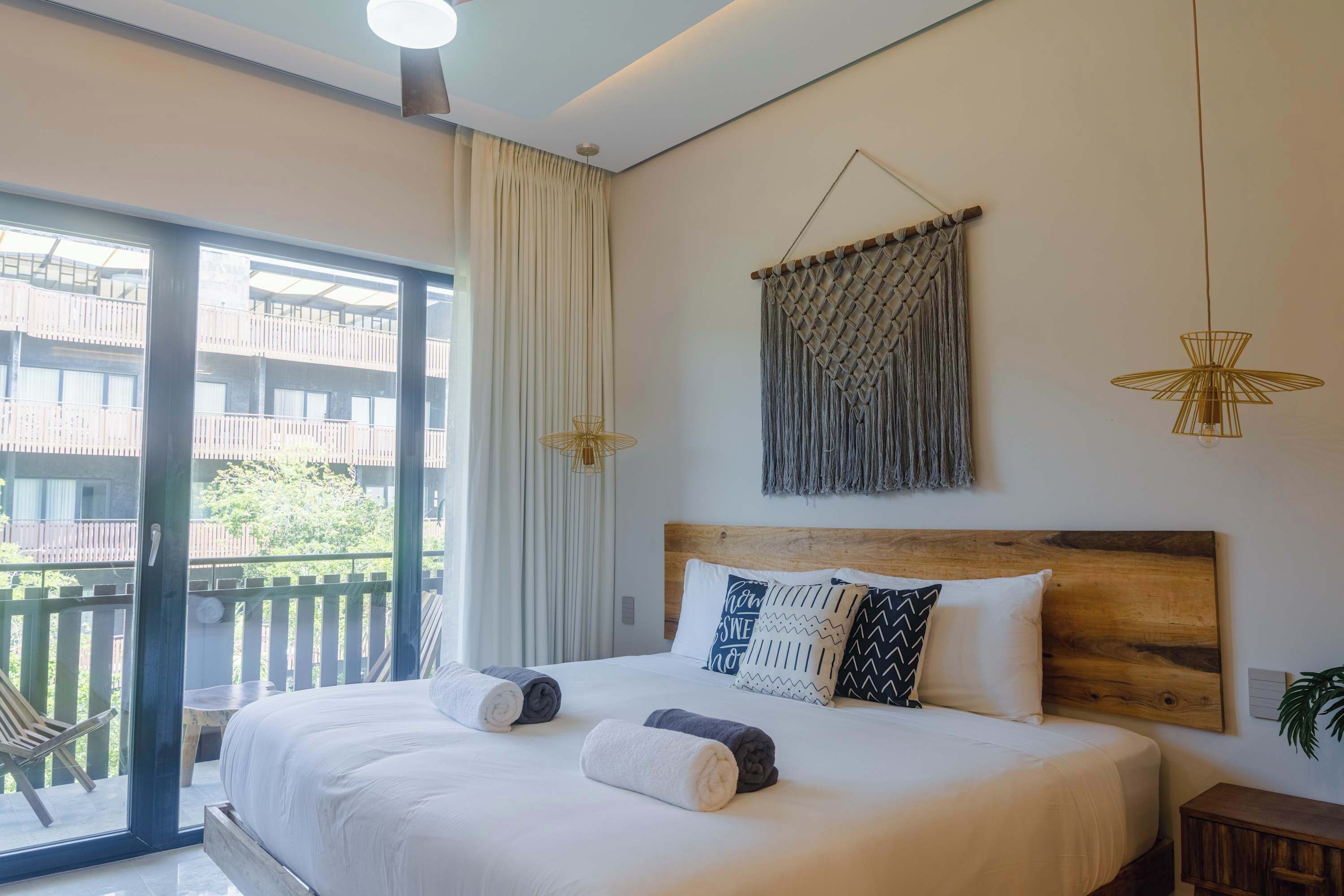 Quarto de hotel com cama, vista para uma varanda e um painel de macramê na parede.